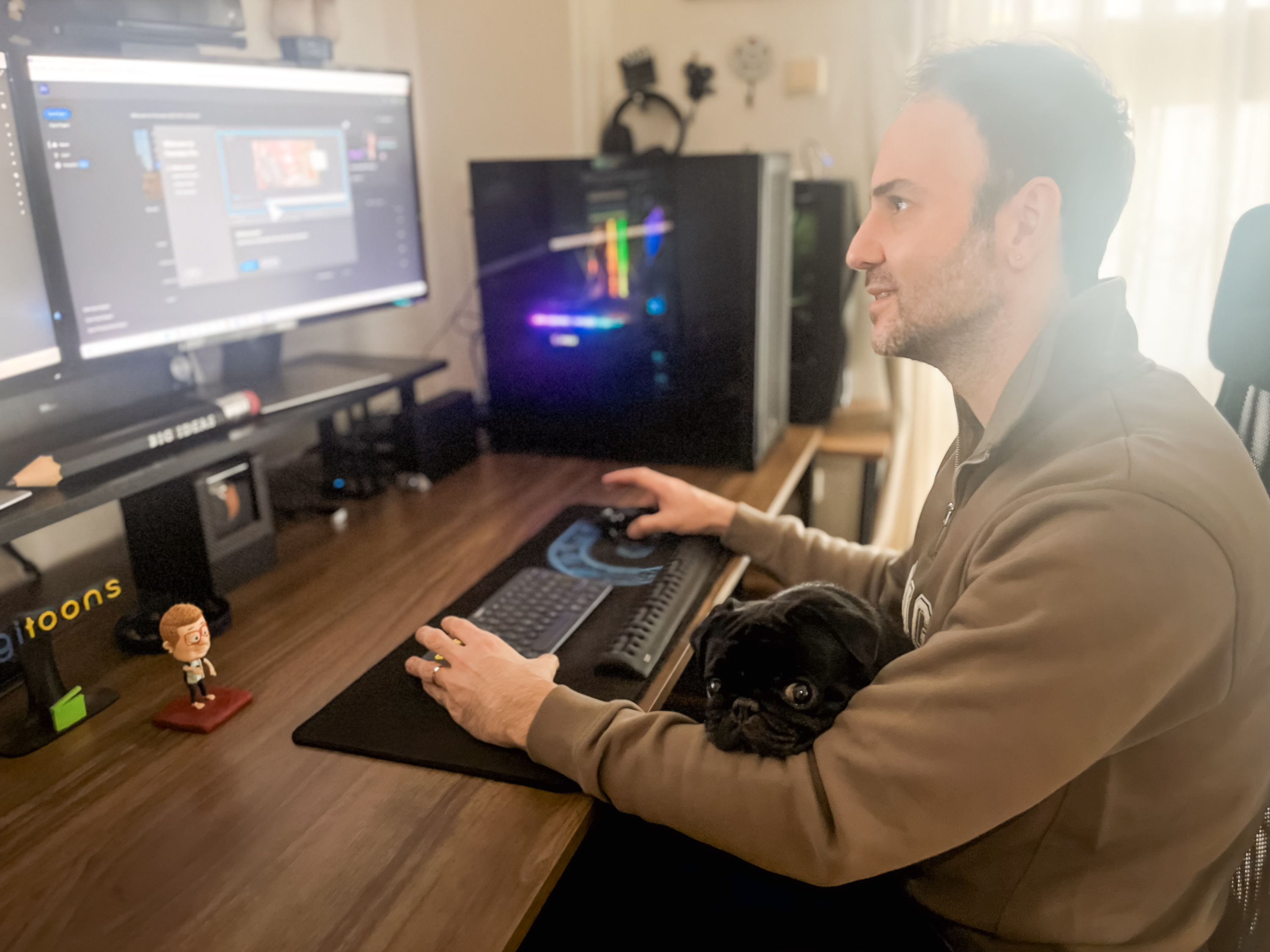 Serhan Yorganci at his desk working, a black pug tucked under his arm, a small Square Heads figure and a Big Ideas pen on the monitor stand beside him
