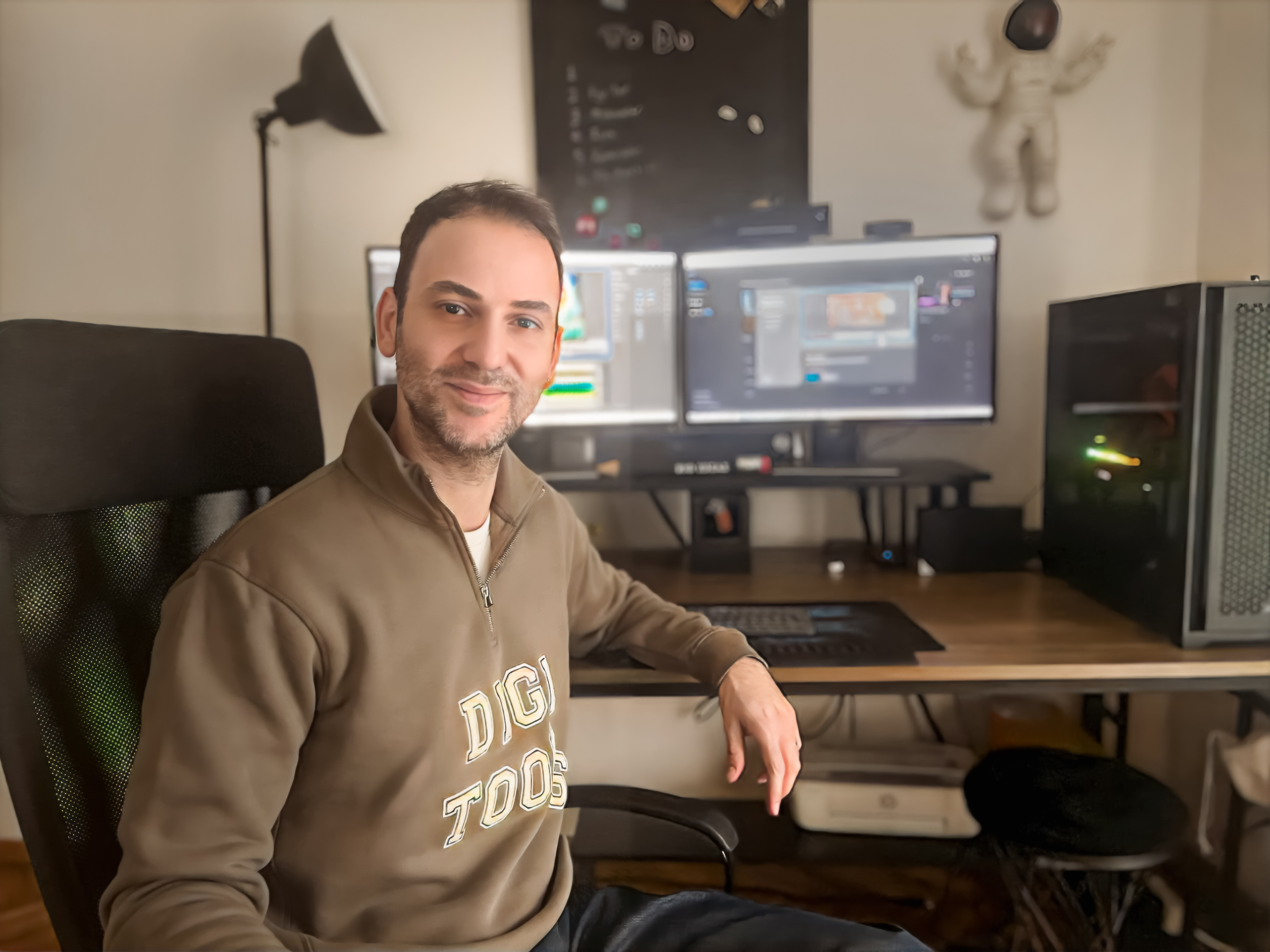 Serhan Yorganci smiling at the camera in his studio, wearing a Digitoons sweatshirt, monitors and workstation behind him
