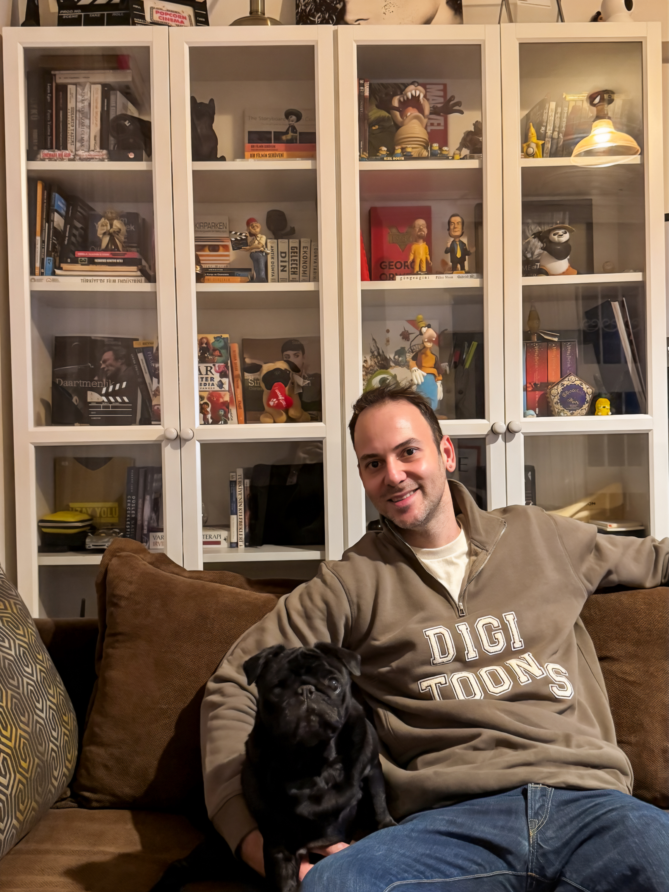 Serhan Yorganci relaxing on a couch with his black pug, a glass cabinet behind him full of film books and figures including Pixar, Kung Fu Panda and Harry Potter